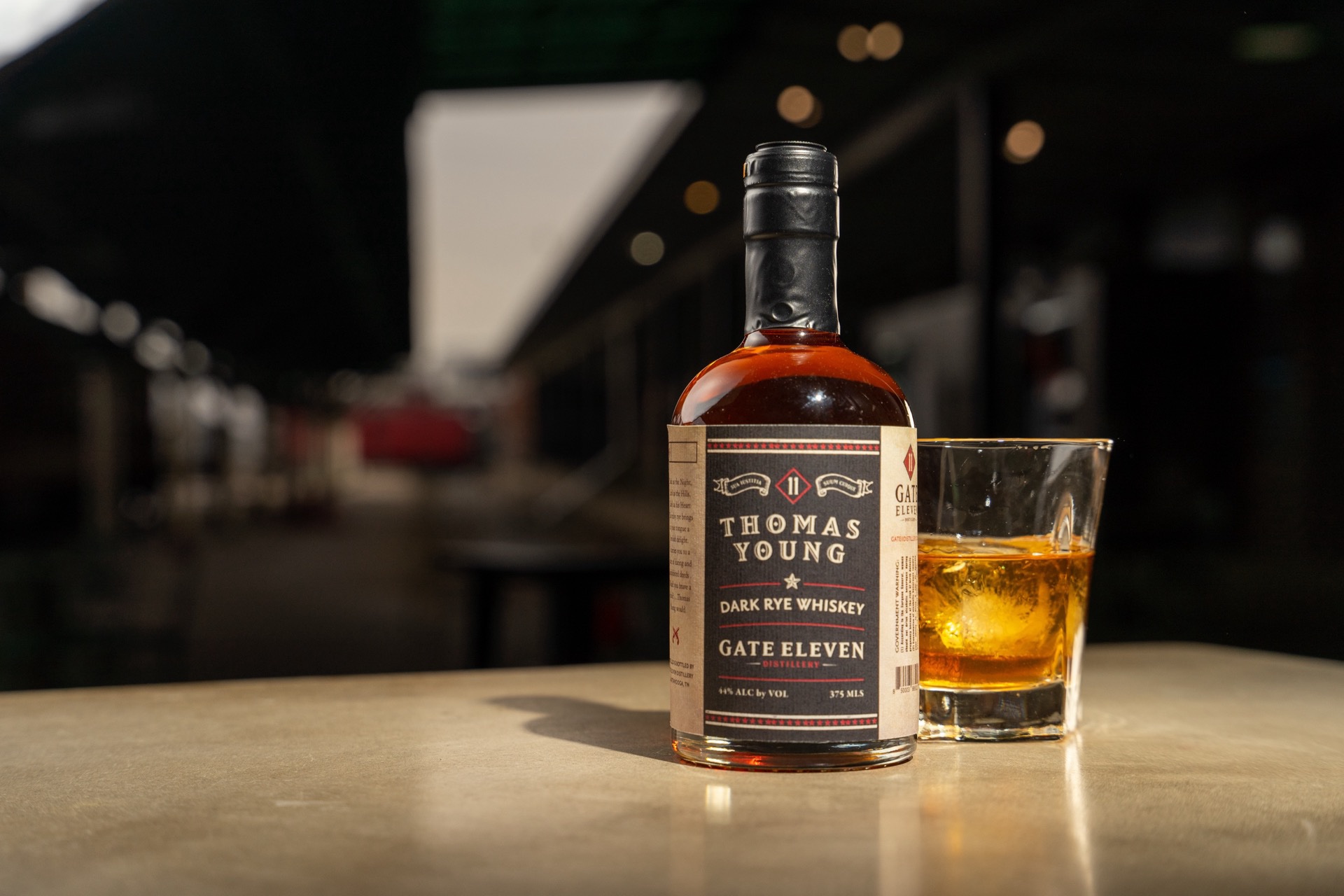 Thomas Young dark rye upright on a sunlit bar top, rocks pour alongside