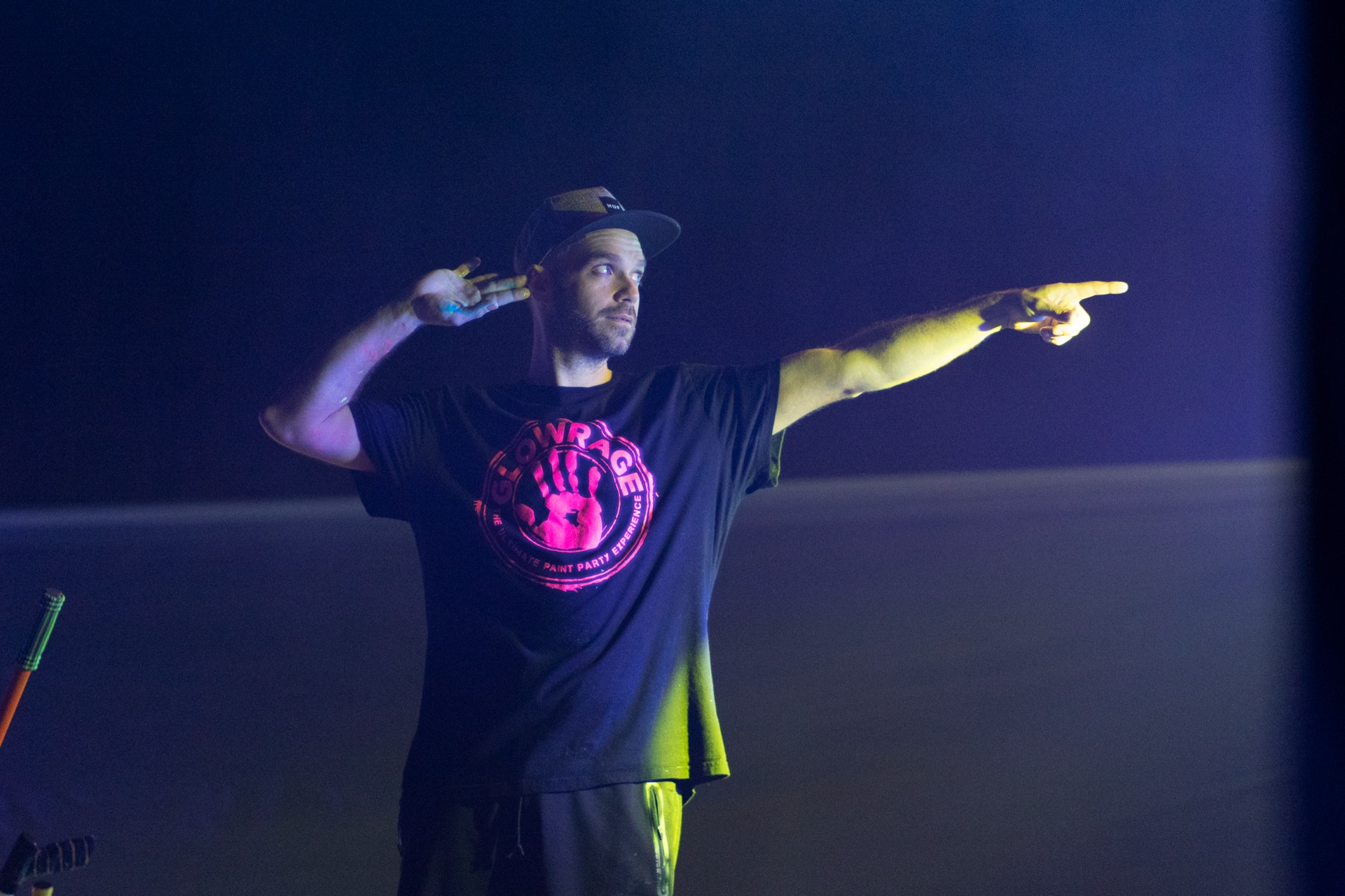 Performer in a GlowRage tee pointing across the dark stage, lit by a single warm beam